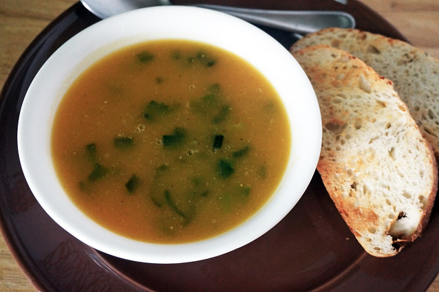 A serving of butternut squash soup with ham and scallions in a small white bowl, dark green scallions and small bubbles punctuating the rich golden broth. In the background, slices of toast and a stainless steel spoon rest on the dark brown plate holding the bowl.