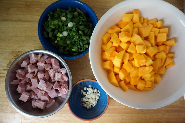 A colorful array of ingredients (orange squash cubes, pink ham, green scallions, ivory garlic) in bowls of various sizes and colors (ivory ceramic, stainless steel, blue ceramic, blue enamel).