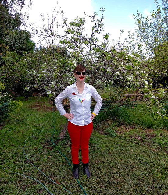 A woman stands in a garden, wearing a white button up shirt and red skinny pants.