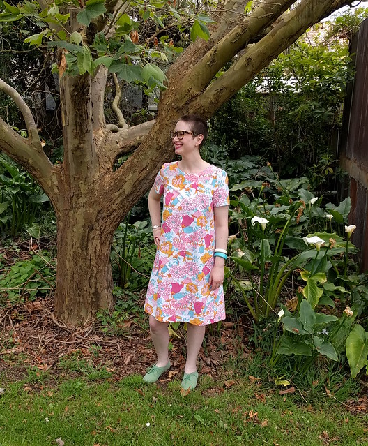 A woman poses in a garden in a retro floral print shift dress and green flock brogues.