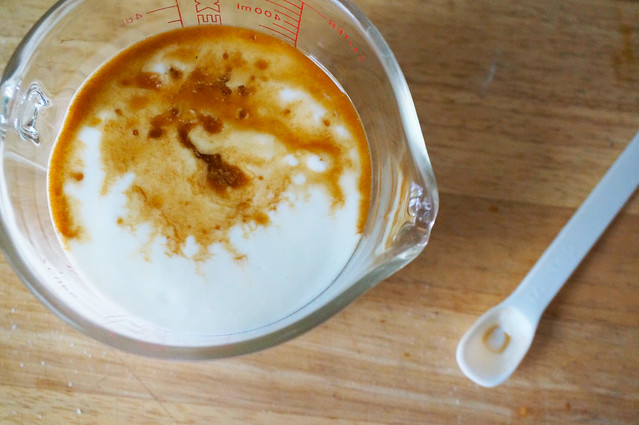 Buttermilk in a glass measuring cup, seen from above, with a small bit of vanilla poured on top. The vanilla doesn't sink in, but instead swirls on top, looking almost like brown clouds in a stark-white sky.