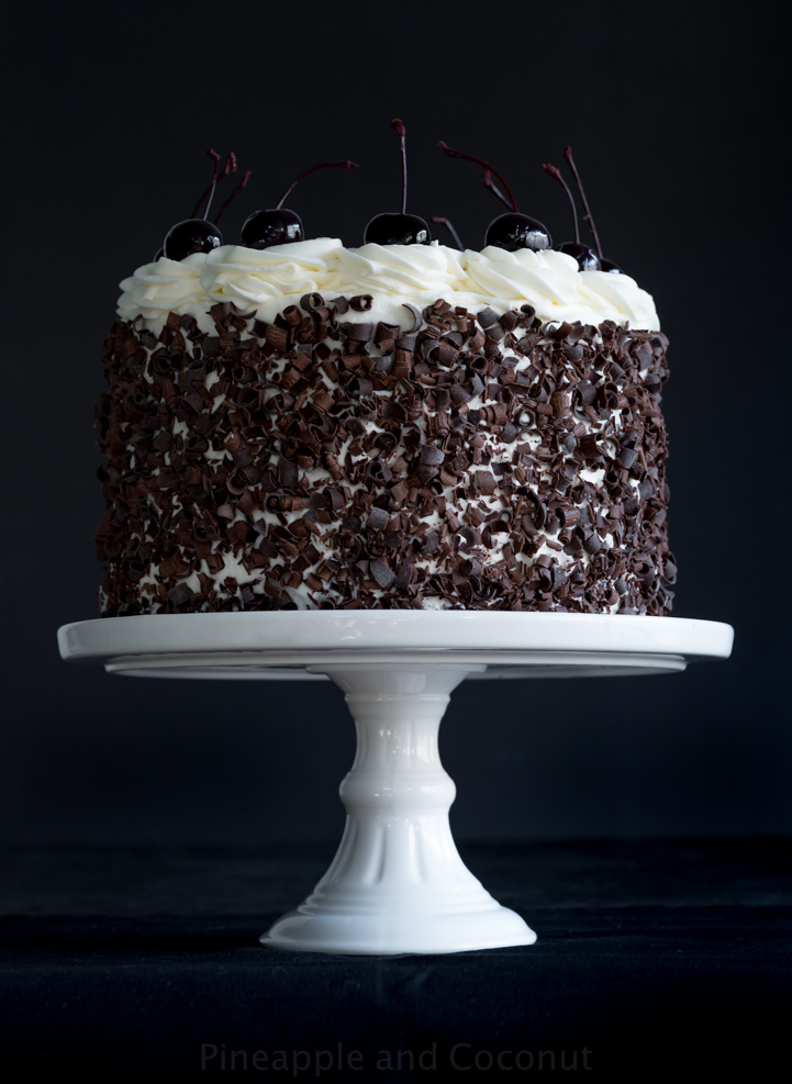 layer cake covered in chocolate curls on a white cake stand, decorated with whipped cream and dark cherries